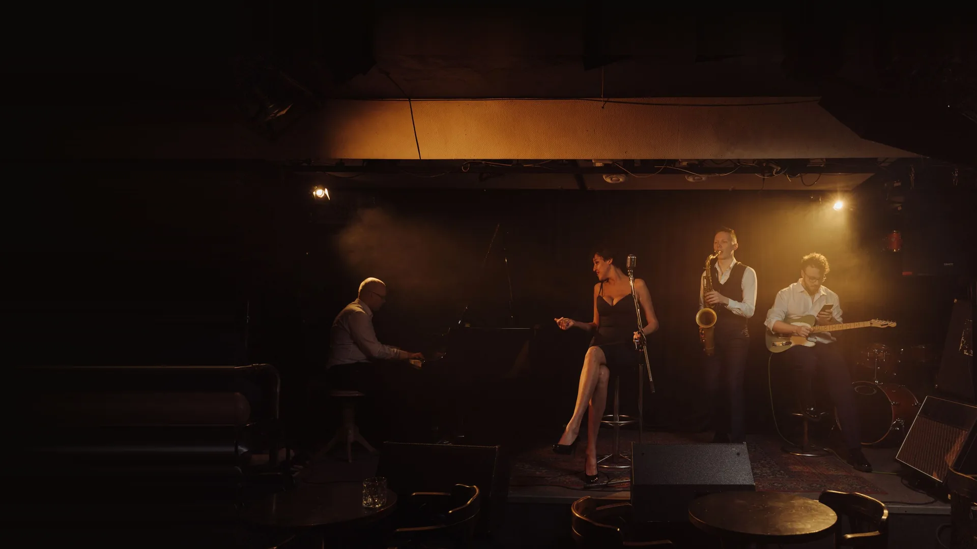 En jazzkvartet på en lille, varmt oplyst klubscene: en pianist ved et flygel, en siddende sanger ved en vintage mikrofon, en saxofonist og en guitarist, med et mørkt publikumsområde i forgrunden.