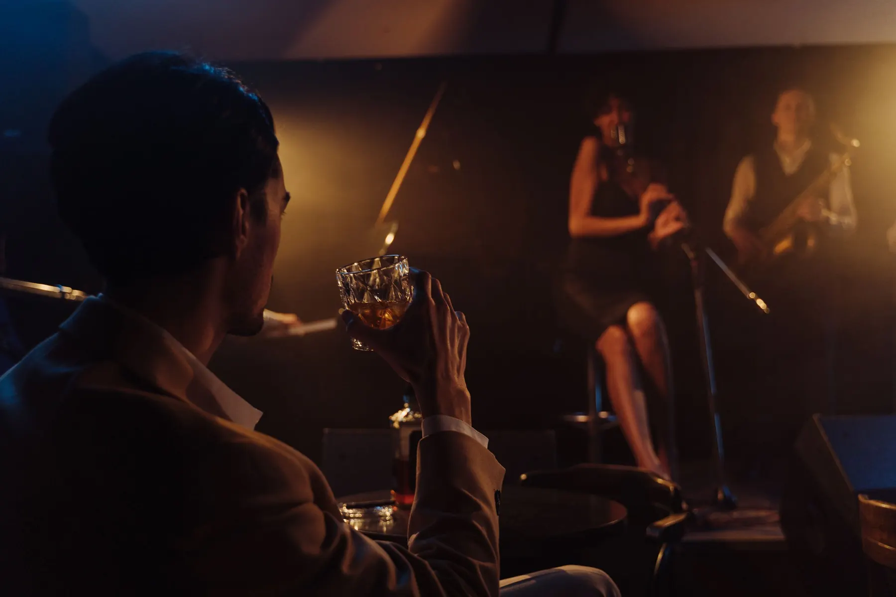 En mand i en brun jakke med et glas whisky i hånden, set bagfra, betragter et jazzband på en oplyst scene bag et lille bord.