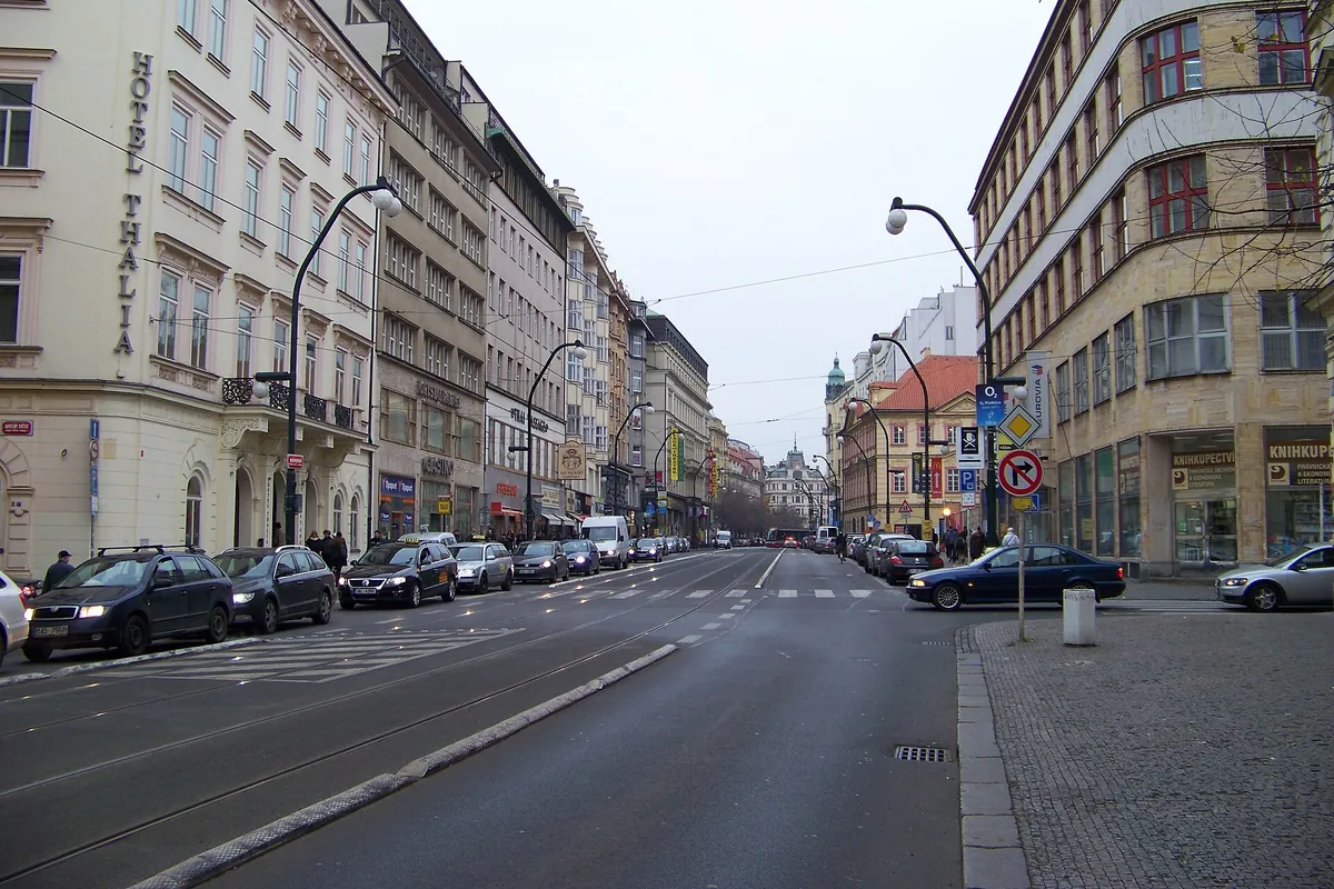 En bred gade i Prag med sporvognsskinner og parkerede biler, kantet af høje bygninger i creme og sten, der strækker sig ud mod horisonten.