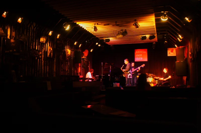 Et jazzband, der spiller på en lille scene oplyst i orange, med et rødt REDUTA JAZZ CLUB-skilt bag musikerne og et mørkt publikumsområde foran.